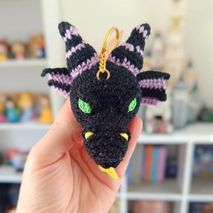 Puede incluir: Un llavero de drag&oacute;n negro tejido a crochet con detalles en morado y amarillo. El drag&oacute;n tiene ojos verdes y una lengua amarilla.