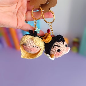 Peut inclure: Un porte-clés en crochet composé de deux personnages : une femme blonde et un homme aux cheveux noirs. Les porte-clés sont attachés à des anneaux dorés et ornés d'un petit charm en forme de fleur bleue. Les porte-clés sont tenus dans une main sur un fond violet. Les étiquettes attachées aux porte-clés indiquent "with love" et "handmade with love".