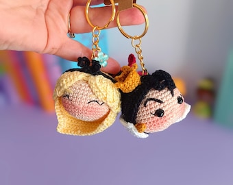May include: A pair of crocheted keychains featuring a blonde woman and a man with black hair. The keychains are attached to gold key rings and have a small blue flower charm. The keychains are held in a hand against a purple background. The tags attached to the keychains read "with love" and "handmade with love".