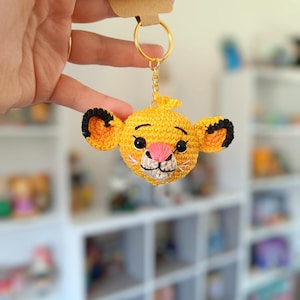 May include: A crocheted lion keyring with black ears and a pink nose. The keyring is attached to a gold chain and ring.