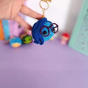 May include: A blue crocheted keychain with a character from the movie Inside Out. The character has blue hair, glasses, and a sad expression.