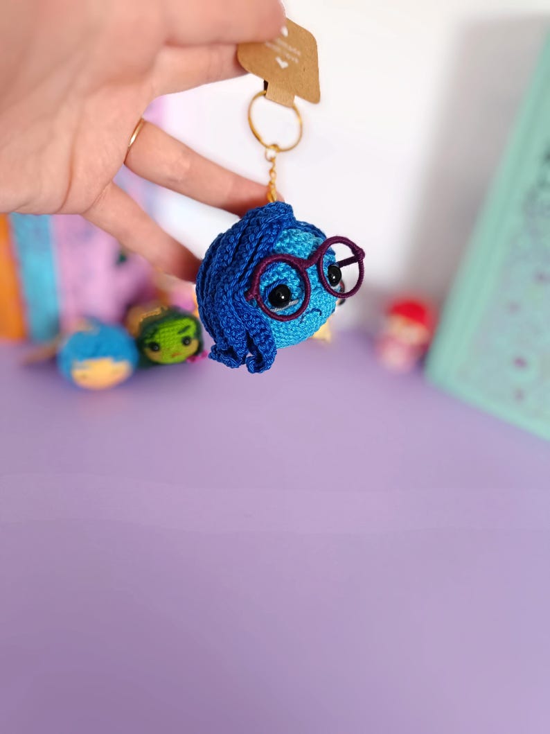 May include: A blue crocheted keychain with a character from the movie Inside Out. The character has blue hair, glasses, and a sad expression.