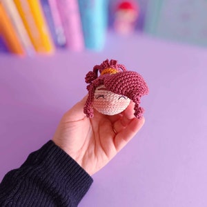 May include: A small crocheted doll with pink hair and a pink and white face. The doll is being held in a hand.