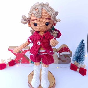 May include: A crocheted doll wearing a red and white outfit with a Christmas tree earring. The doll has blonde hair and is standing on a wooden base.
