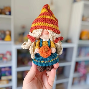Op de afbeelding: Een gehaakte gnoompup met een rood-gele gestreepte muts, een blauw-groen jurkje en een kleine oranje pompoen in de hand.