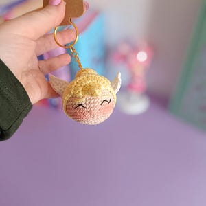 Peut inclure: Un porte-clés en crochet avec un visage jaune et rose, de petites ailes et un anneau doré. Le porte-clés est suspendu à une main.