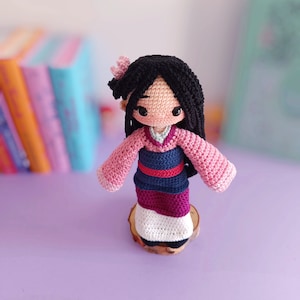 Peut inclure: Une poupée en crochet représentant une jeune femme aux cheveux noirs, vêtue d'une robe rose, bleue et blanche. La poupée est posée sur un socle en bois.