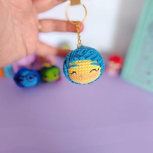 May include: A blue and yellow crocheted keychain with a smiling face. The keychain is attached to a gold key ring.