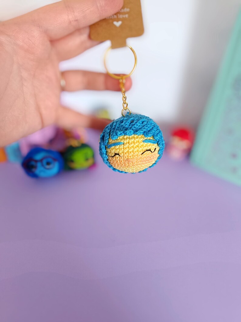 May include: A blue and yellow crocheted keychain with a smiling face. The keychain is attached to a gold key ring.