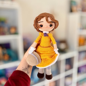 May include: A handmade crochet doll wearing a yellow dress with white trim, a purple bow, and a matching hat. The doll has brown hair and is holding a small, round hat. The doll is approximately 8 inches tall.