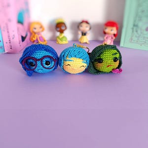 May include: Three crocheted character keychains, each with a different facial expression. The first keychain is blue with a sad expression and glasses. The second keychain is yellow with a happy expression. The third keychain is green with an angry expression.
