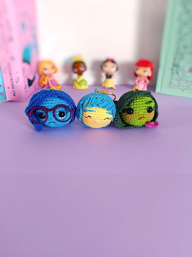 May include: Three crocheted character keychains, each with a different facial expression. The first keychain is blue with a sad expression and glasses. The second keychain is yellow with a happy expression. The third keychain is green with an angry expression.
