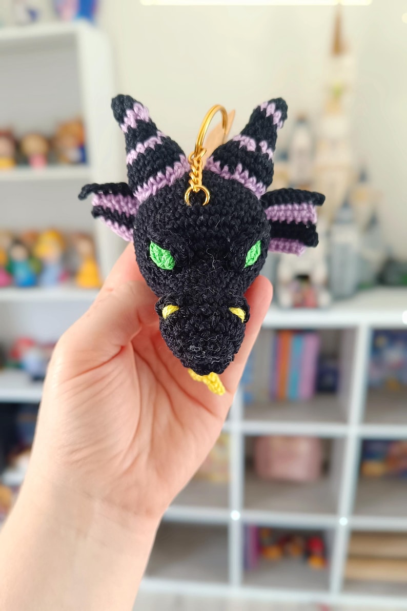 Puede incluir: Un llavero de drag&oacute;n negro tejido a crochet con detalles en morado y amarillo. El drag&oacute;n tiene ojos verdes y una lengua amarilla.