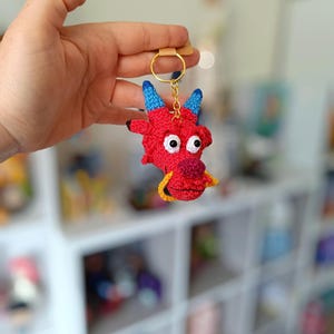 May include: A handmade red crochet dragon keychain with blue horns, white eyes, and a yellow accent. The keychain has a gold ring and a small leather strap for attachment. This whimsical accessory is perfect for adding a touch of fantasy to your keys or bag.