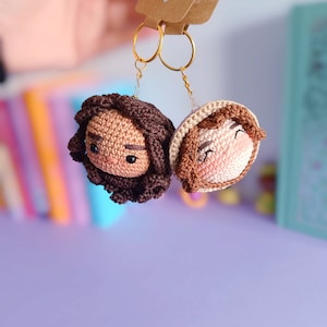 May include: Two crocheted keychains, one with brown hair and the other with light brown hair, are attached to gold key rings. The keychains are hanging from a hand. A small brown tag with the text "Handmade with love" is attached to the key rings.