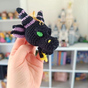 Puede incluir: Un llavero de drag&oacute;n de ganchillo negro y morado con ojos verdes y una estrella amarilla.