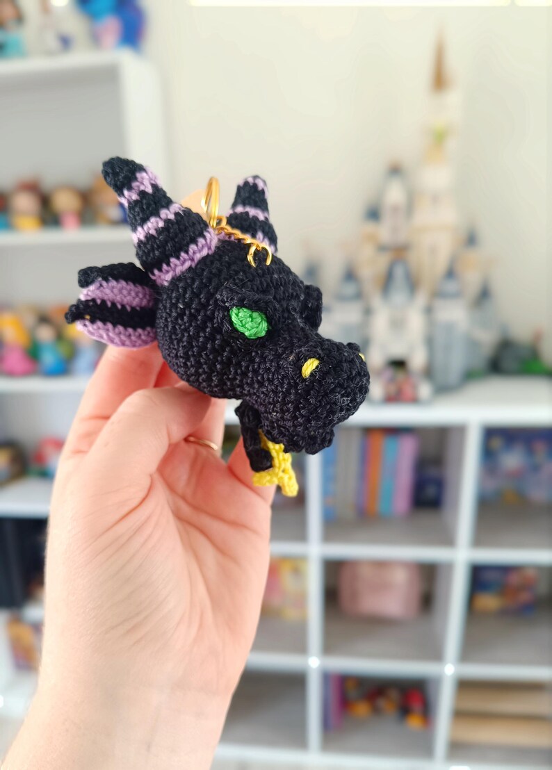 Puede incluir: Un llavero de drag&oacute;n de ganchillo negro y morado con ojos verdes y una estrella amarilla.