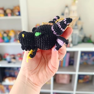 Puede incluir: Un llavero de drag&oacute;n negro tejido a crochet con ojos verdes y detalles morados. El drag&oacute;n est&aacute; sosteniendo una cola amarilla en su boca.