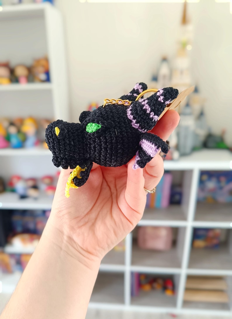 Puede incluir: Un llavero de drag&oacute;n negro tejido a crochet con ojos verdes y detalles morados. El drag&oacute;n est&aacute; sosteniendo una cola amarilla en su boca.