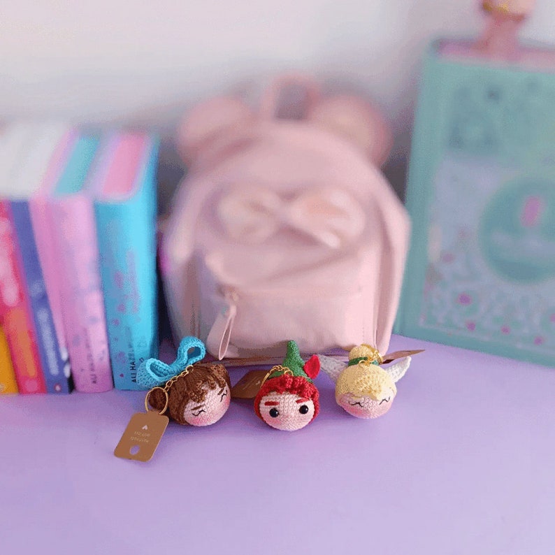 May include: Three crocheted character keychains with a pink backpack in the background. The keychains are a blue haired girl, a red haired boy with a green hat, and a blonde girl with wings.