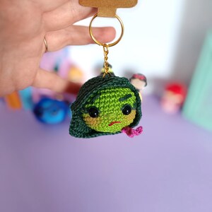 May include: Green crocheted keychain with a cartoon face and a pink bow. The keychain is attached to a gold key ring. The tag says "Handmade with love".