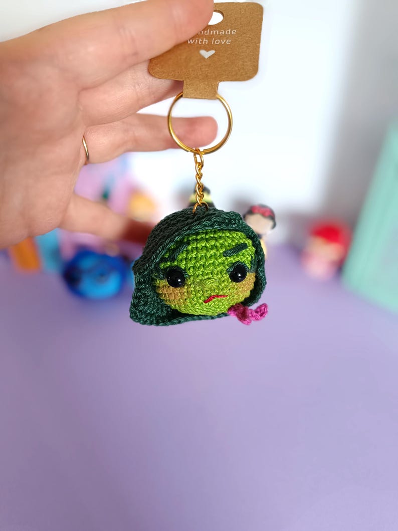 May include: Green crocheted keychain with a cartoon face and a pink bow. The keychain is attached to a gold key ring. The tag says "Handmade with love".