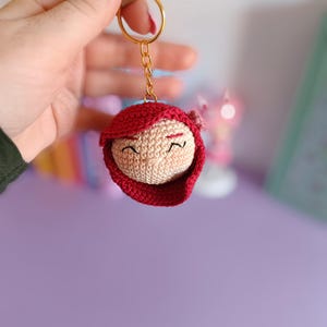 May include: A crocheted keychain featuring a red-haired girl with a pink flower in her hair. The keychain is attached to a gold chain and ring.