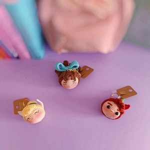 May include: Three crocheted doll keychains on a purple surface. The dolls have brown, blonde, and red hair and are wearing different outfits. Each doll has a brown paper tag attached to it.
