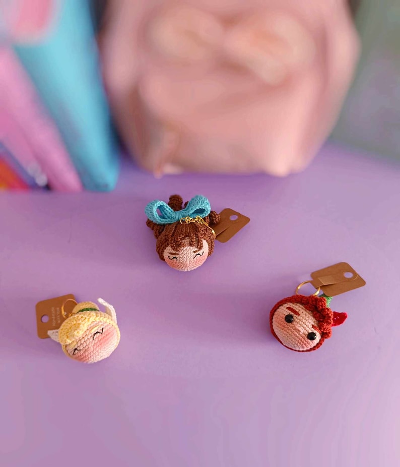 May include: Three crocheted doll keychains on a purple surface. The dolls have brown, blonde, and red hair and are wearing different outfits. Each doll has a brown paper tag attached to it.