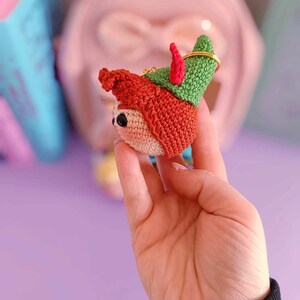 May include: A crocheted elf ornament with a red hat, green pointed hat, and a gold key ring. The elf has a small, round face with black eyes and a pink nose.