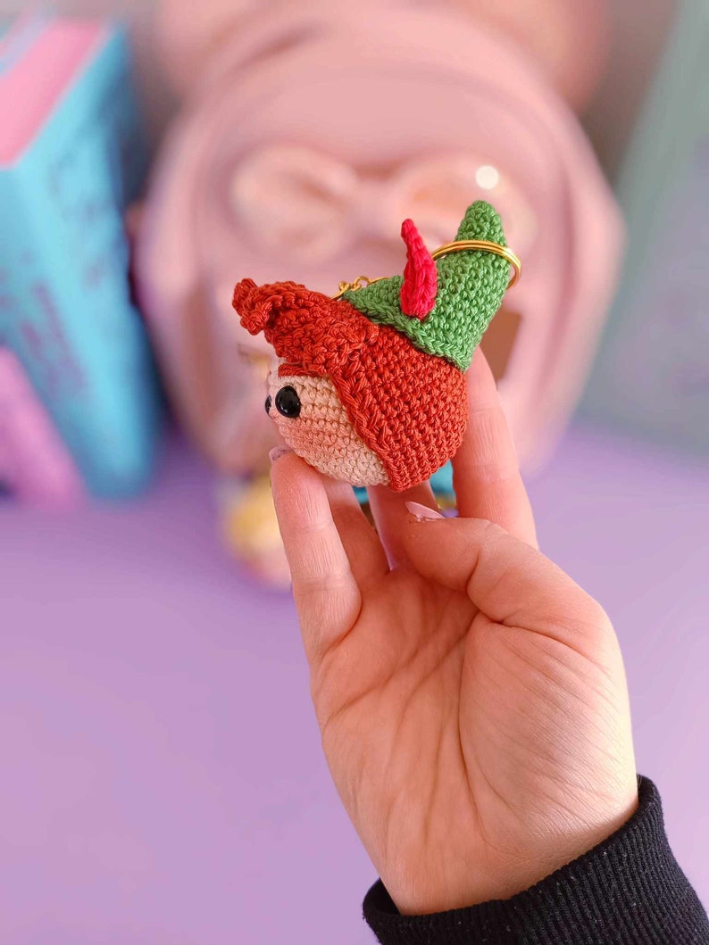 May include: A crocheted elf ornament with a red hat, green pointed hat, and a gold key ring. The elf has a small, round face with black eyes and a pink nose.