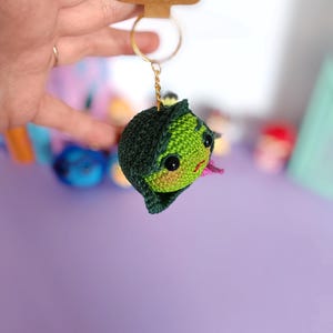 May include: Green crocheted keychain with a pink mouth and black eyes. The keychain is attached to a gold key ring. The tag says "Handmade with love".