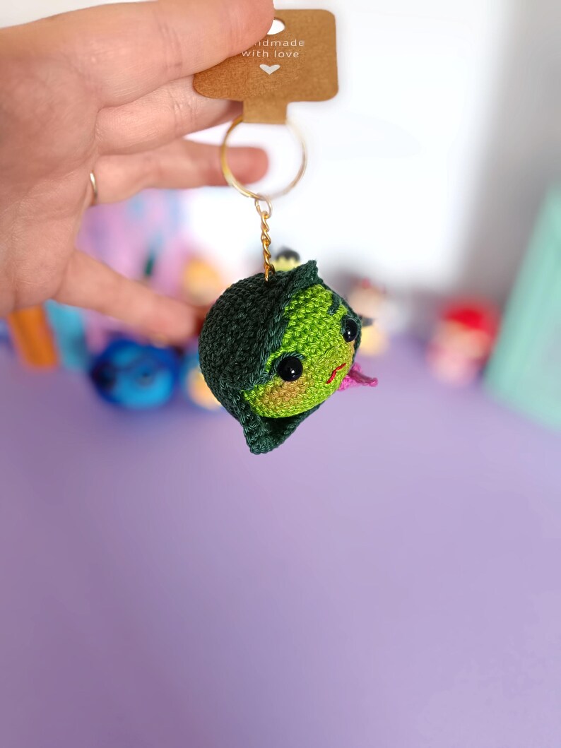 May include: Green crocheted keychain with a pink mouth and black eyes. The keychain is attached to a gold key ring. The tag says "Handmade with love".