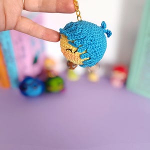 May include: A crocheted keychain featuring a smiling face with blue hair. The keychain is attached to a gold key ring.