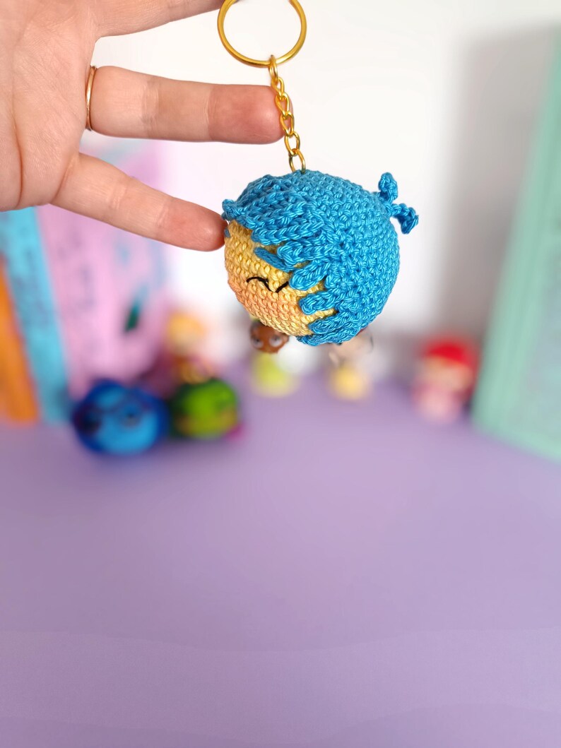 May include: A crocheted keychain featuring a smiling face with blue hair. The keychain is attached to a gold key ring.
