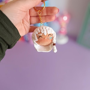 May include: A crocheted keychain with a white face and brown hair. The keychain has a gold chain and a tag that says "made with love".