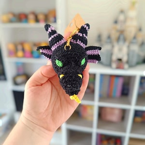 Puede incluir: Un llavero de drag&oacute;n negro tejido a crochet con rayas moradas y negras, ojos verdes y cola amarilla.