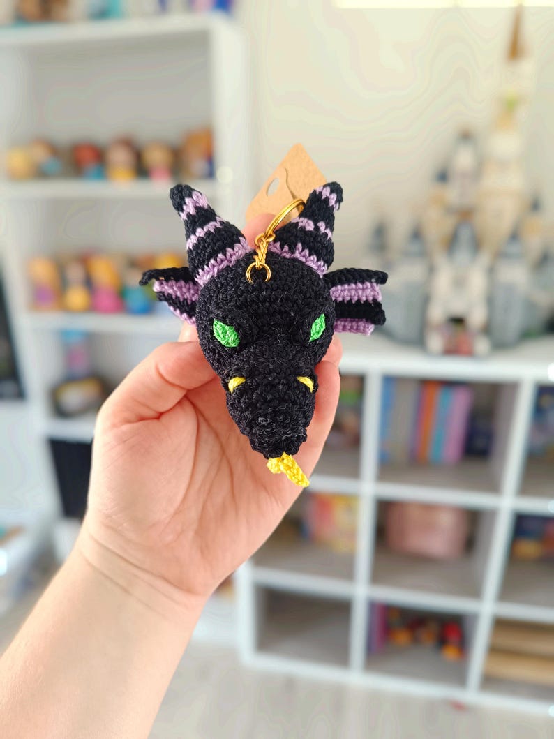 Puede incluir: Un llavero de drag&oacute;n negro tejido a crochet con rayas moradas y negras, ojos verdes y cola amarilla.