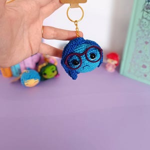 May include: Blue crocheted keychain with a character from the movie Inside Out. The character has glasses and a sad expression. The keychain is attached to a gold key ring with a brown tag that says "handmade with love".