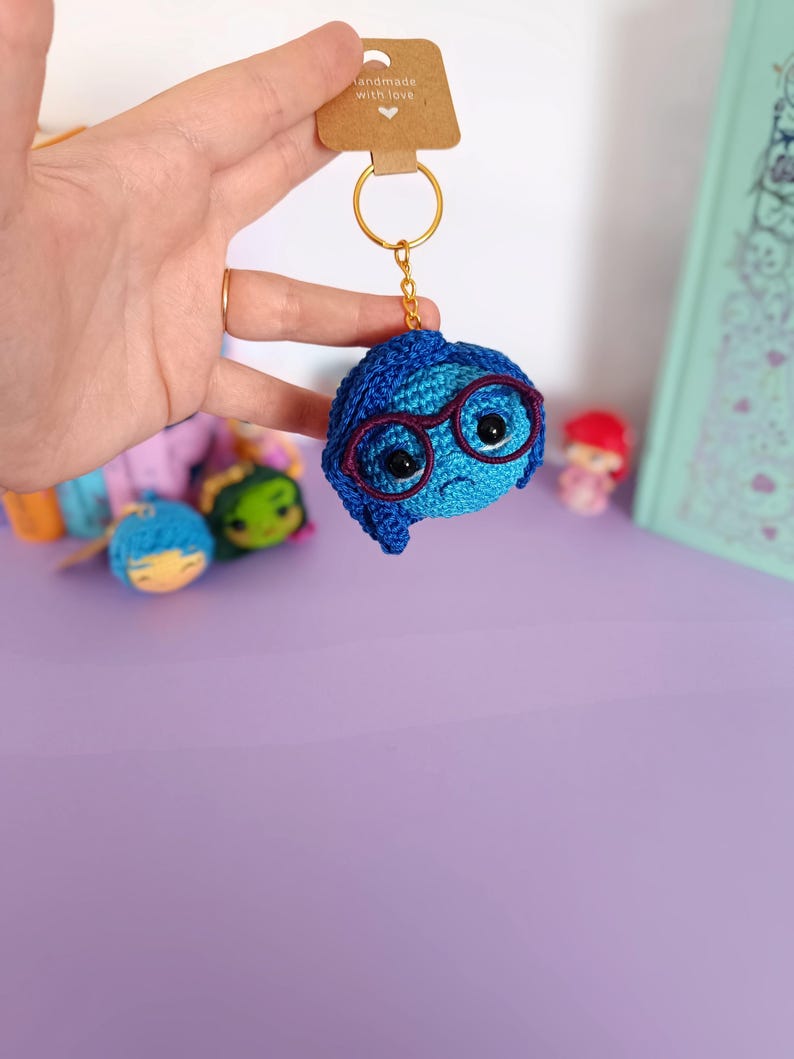 May include: Blue crocheted keychain with a character from the movie Inside Out. The character has glasses and a sad expression. The keychain is attached to a gold key ring with a brown tag that says "handmade with love".