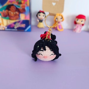 May include: A crocheted keychain featuring a cartoon character with black hair, pink cheeks, and a red bow. The keychain is attached to a gold key ring.