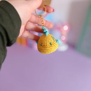 Peut inclure: Un porte-clés en crochet jaune en forme de poisson avec des accents bleus et verts. Le porte-clés est attaché à un anneau de clé en or.