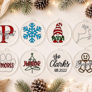 May include: Eight clear circular acrylic ornaments featuring various holiday designs including a red letter P with the name Payton, a blue snowflake, a gnome with the name Nana, a reindeer, a paw print with S'mores, a tree with Junior, the text the Clarks EST 2022, and a gingerbread man with Brayden.