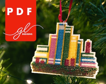 May include: A colorful cross-stitch ornament shaped like a stack of books. The ornament is decorated with red, pink, yellow, green, blue, and white thread. The ornament is hanging on a Christmas tree.