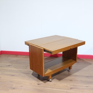 May include: A mid-century modern wooden tea cart with a rectangular top and a lower shelf. The cart has a pull-out extension, and it sits on small caster wheels. The wood has a warm, light brown finish.