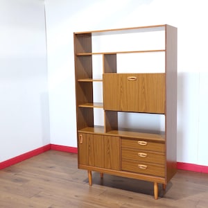 May include: A tall wooden room divider shelving unit with a light brown wood grain finish. It features open shelves, a central drop-down cabinet door, a lower cabinet door on the left, and three stacked drawers on the right with light handles.