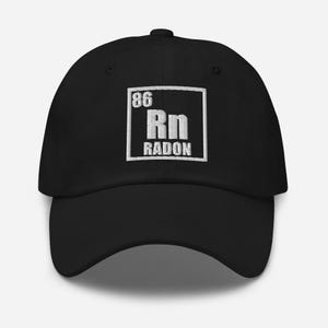 May include: A black baseball cap featuring an embroidered white square patch on the front. Inside the square is the chemical element symbol for Radon, displaying the number 86, the letters Rn, and the word RADON in a white stitched font.
