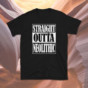 Può includere: T-shirt nero con la scritta bianca "STRAIGHT OUTTA NEOLITHIC" in un carattere usurato, disposta su tre righe all'interno di un rettangolo bianco. La maglietta ha un girocollo e maniche corte.