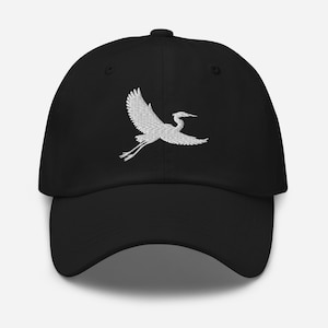 Op de afbeelding: Een zwarte baseballpet met een wit geborduurd ontwerp van een vliegende kraanvogel. De pet heeft een gestructureerde kroon, een gebogen klep en oogjes voor ventilatie. De vogel is afgebeeld met gespreide vleugels en uitgestrekte poten.