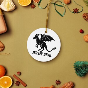 May include: A round white ceramic ornament featuring a black silhouette graphic of the Jersey Devil with wings and horns. The text JERSEY DEVIL is printed in black capital letters below the creature. A gold string is attached for hanging.